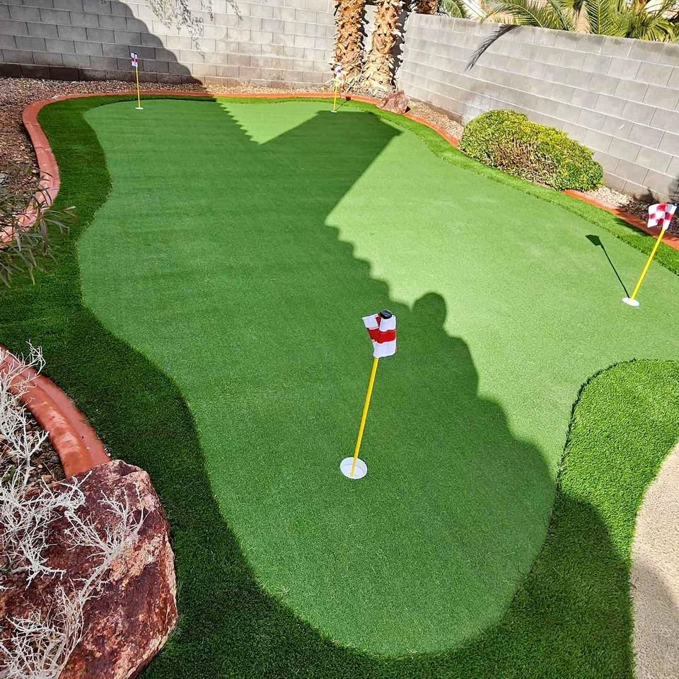 A backyard putting green by Flagstaff Turf and Hardscape features three holes with red and white flags, bordered by rocks, plants, and a stone wall. Palm trees cast shadows across the lush green, creating a relaxing outdoor escape.