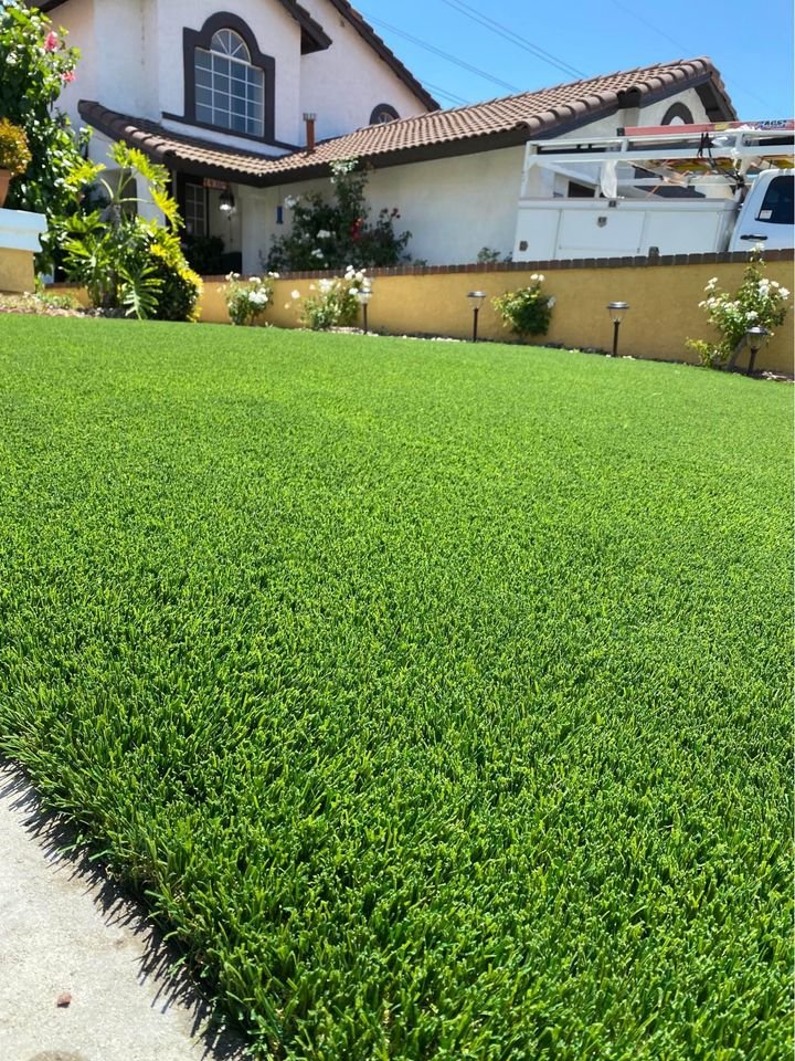 A neatly maintained green lawn in front of a suburban house with a tiled roof, large windows, and garden showcases the expert touch of Flagstaff Turf and Hardscape, with a white work truck parked nearby on a sunny day.