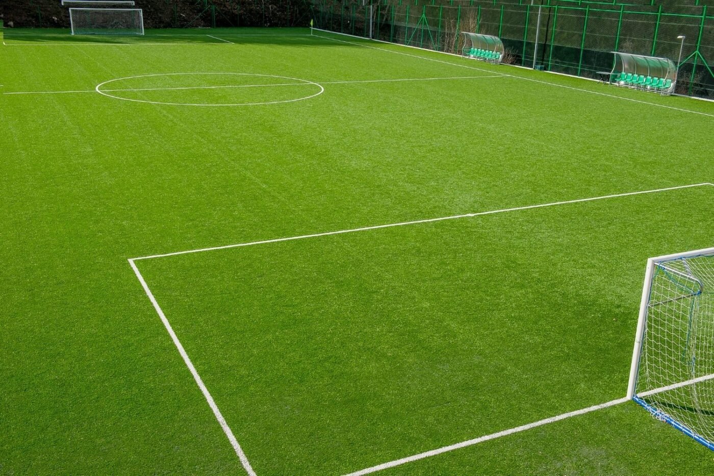 A well-maintained, empty soccer field with bright green Sports Turf, crisp white boundary lines, two goals, benches, and a green fence along one side.