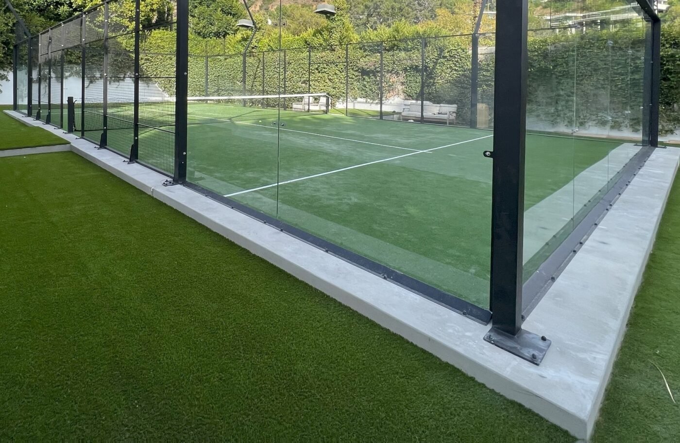 A modern outdoor padel court surrounded by glass walls and metal posts, featuring Sports Turf outside the court and greenery in the background.