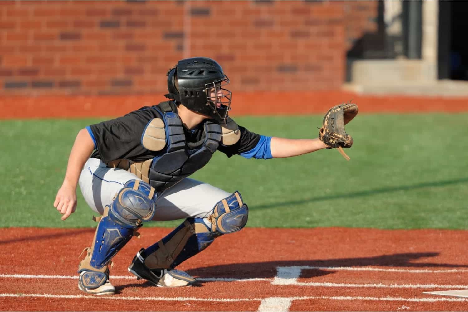 A baseball catcher wearing protective gear crouches behind home plate, extending his gloved hand to catch a pitch on sunlit sports turf with a brick wall in the background.
