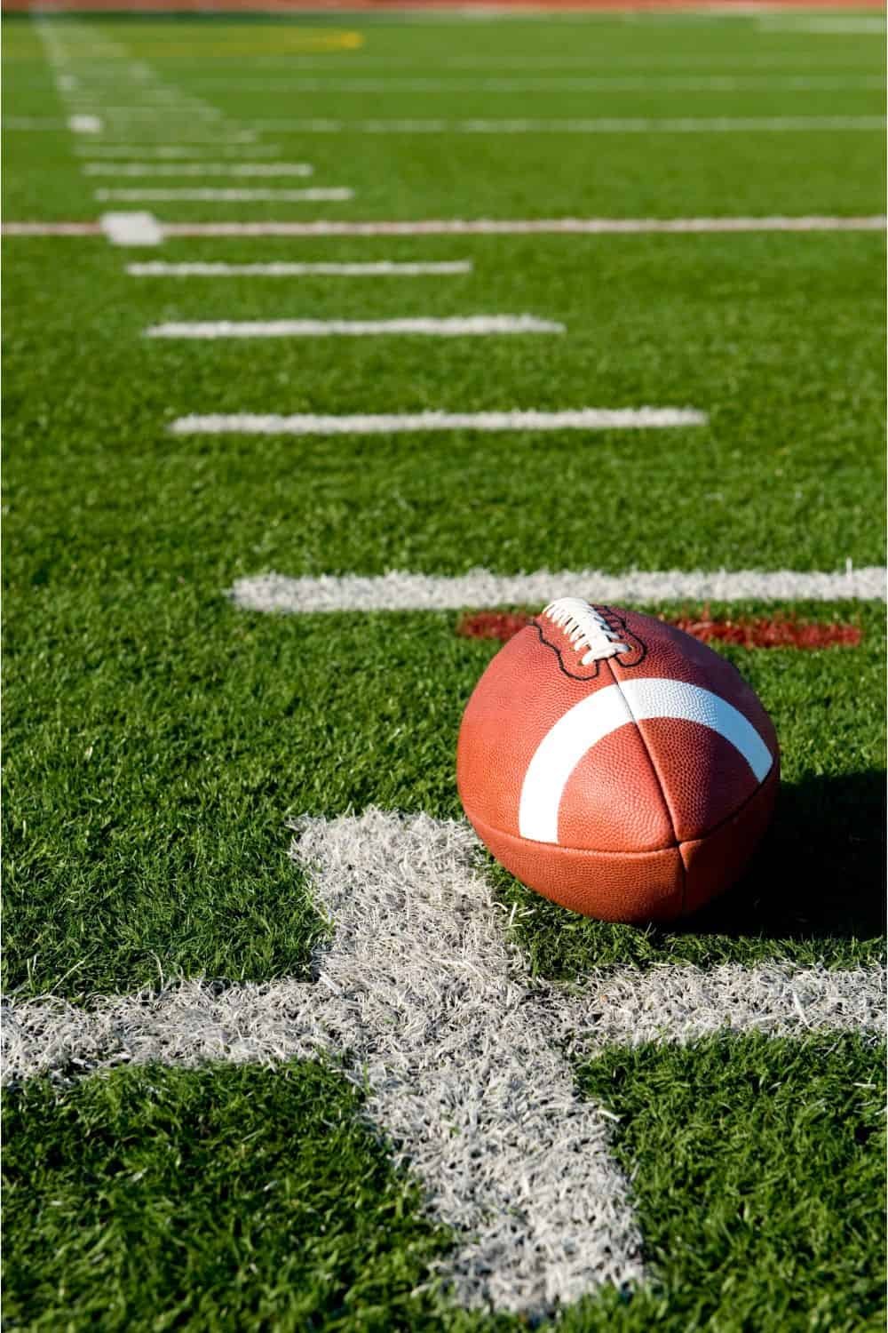 A football rests on the green sports turf of a field, positioned near a white yard line with other yard markings visible in the background.