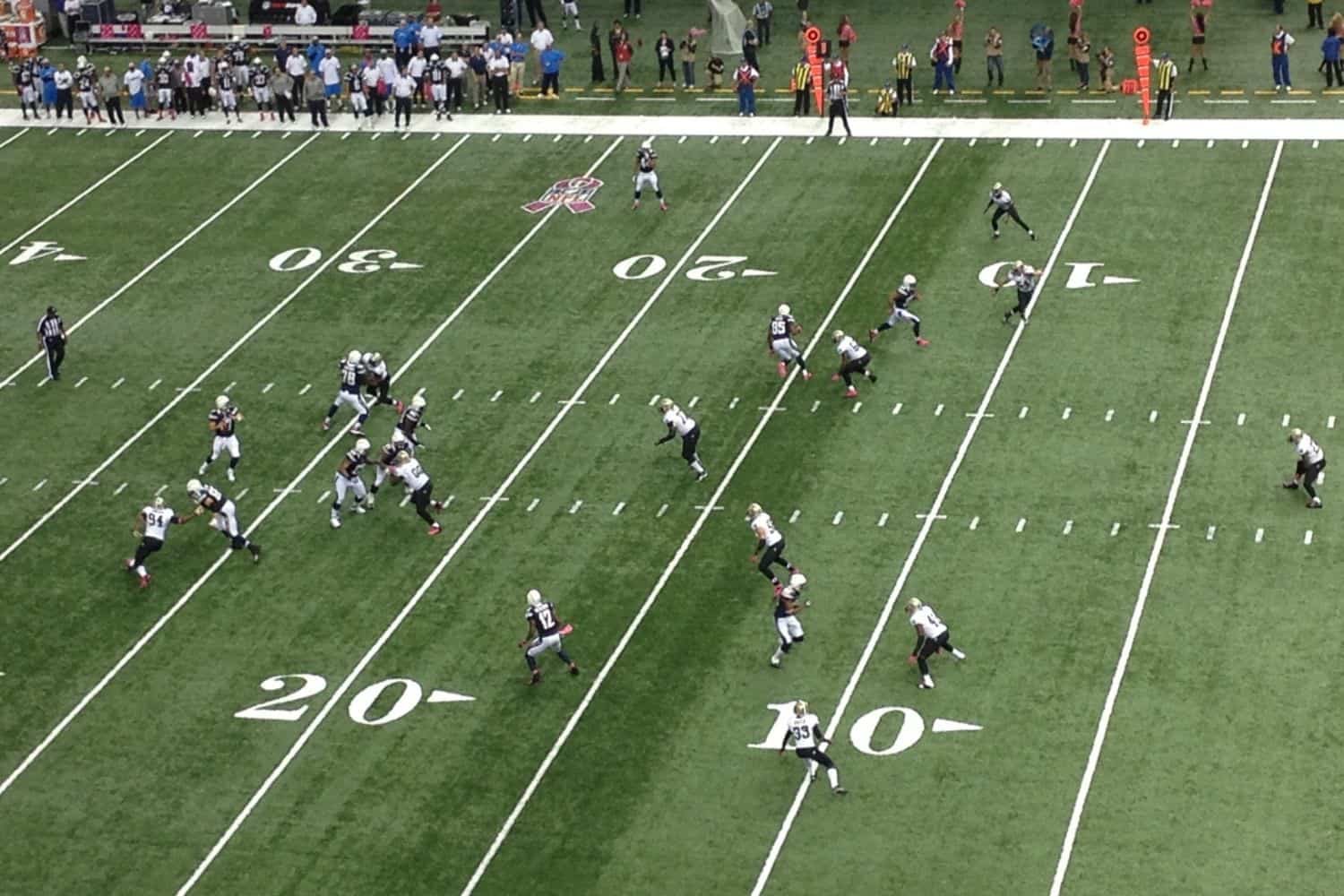 Aerial view of an American football game in progress on vibrant sports turf, with players in black and white uniforms spread near the 10- and 20-yard lines, and spectators visible along the sidelines.
