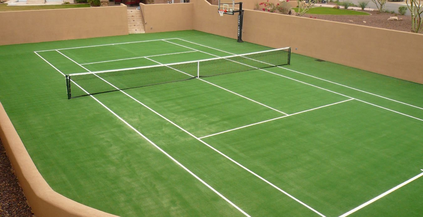 A green sports turf tennis court with white boundary lines is surrounded by tan walls. There is a net in the center and a basketball hoop at one end. Steps lead up from the court, with desert landscaping visible outside the walls.