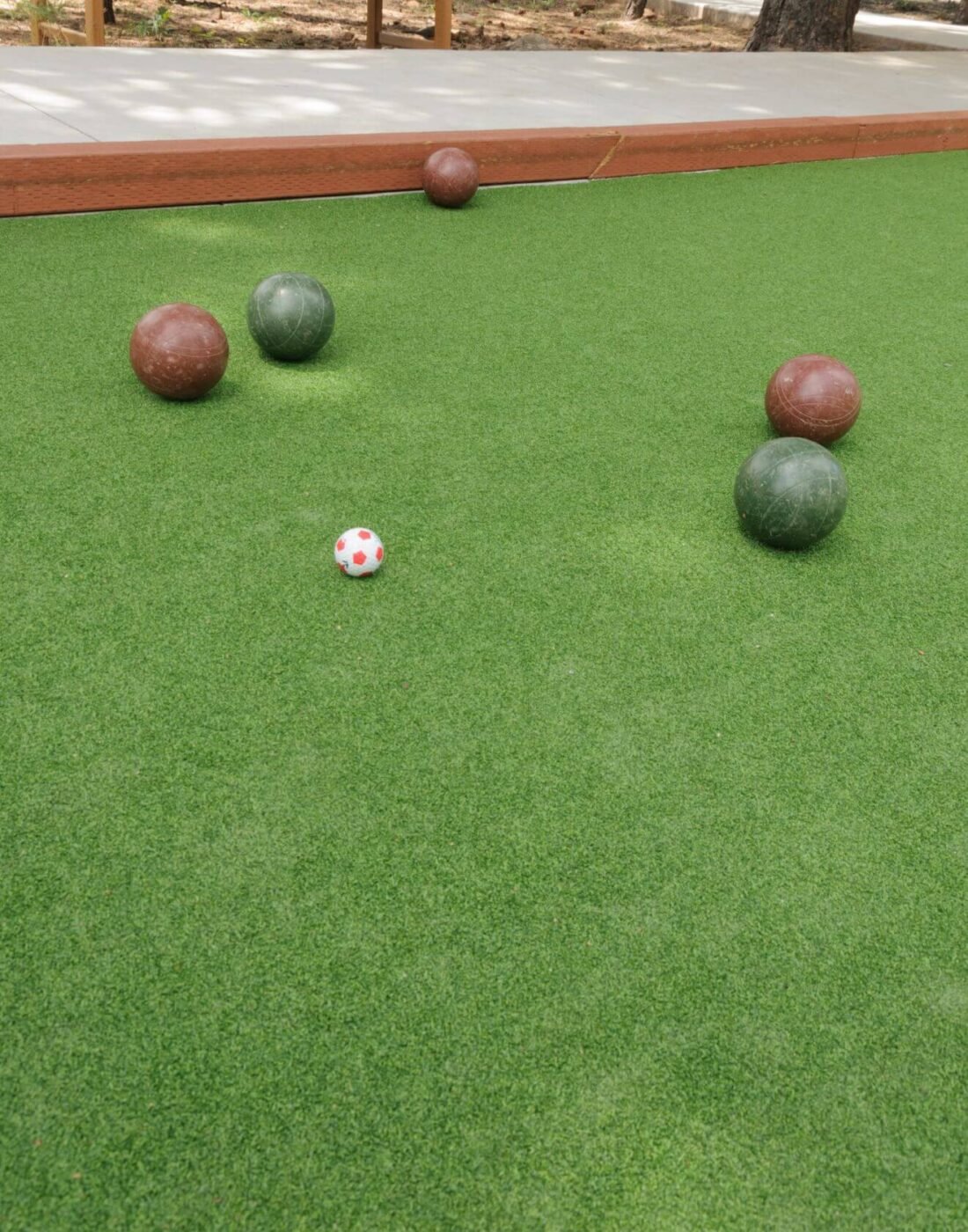 Four bocce balls and a small white-and-red pallino ball are spread out on green sports turf, with a wooden border edging the artificial bocce court.
