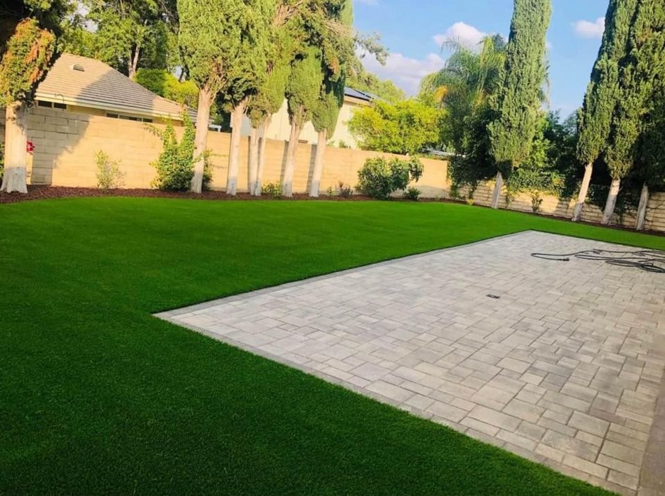 A backyard with a neatly trimmed green lawn, bordered by tall trees and a stone wall. A large rectangular paved area, showcasing expert paver installation, sits in the foreground with a coiled hose on it. The scene is bright and sunny.