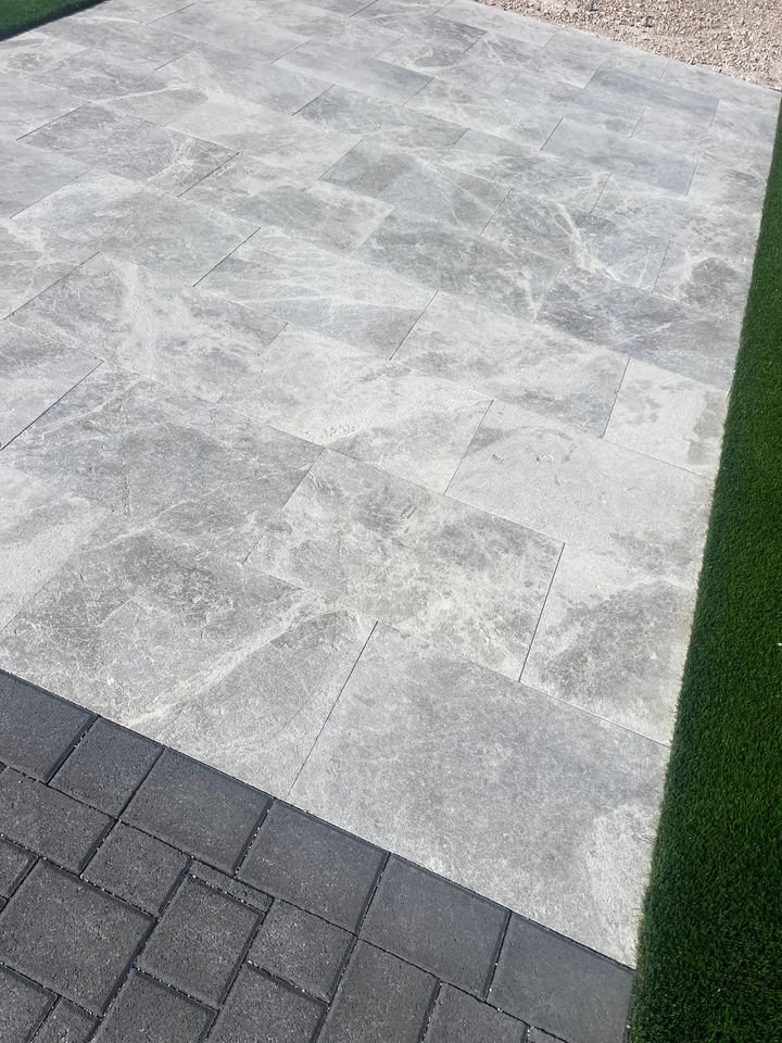 Gray stone patio tiles bordered by dark pavers at the base create a polished look, with artificial green grass lining the sides; professional paver installation ensures lasting beauty, while gravel accents the upper right corner.