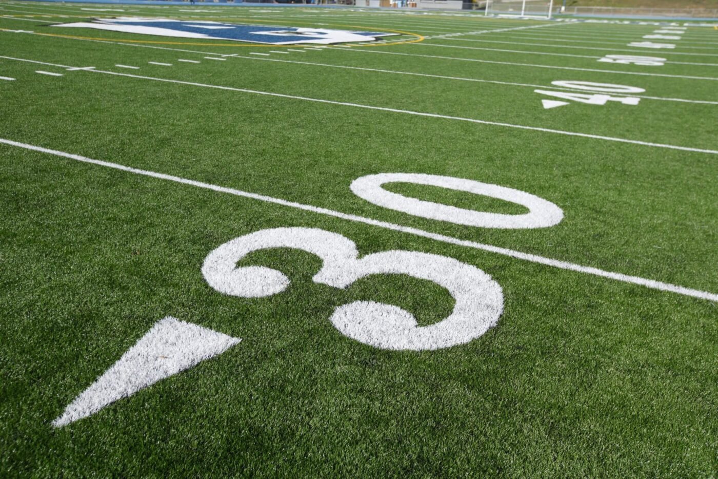 Close-up view of a football field with white yard lines and the number 30 clearly marked on green artificial turf by Flagstaff Turf and Hardscape. A painted logo is visible in the background near the center of the field.